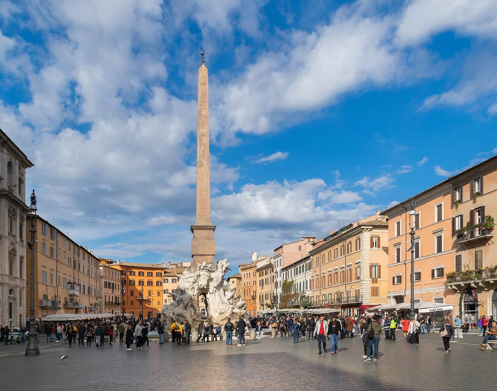 Piazza Navona a Roma con la Fontana dei Quattro Fiumi e l'obelisco al centro, palazzi color ocra ai lati e passanti sotto un cielo azzurro con nuvole