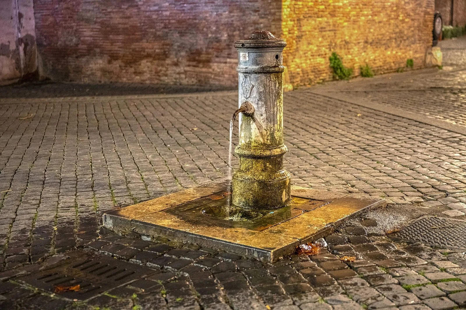 Nasone, la tipica fontanella pubblica romana in ghisa, con l'acqua che scorre su una piazzetta di sampietrini illuminata la sera